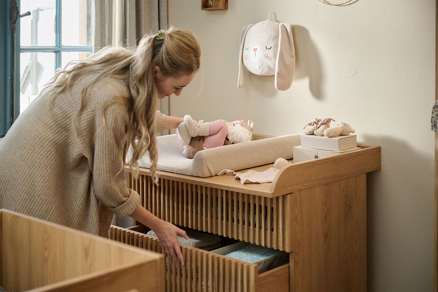 Une femme prépare un bébé sur une table à langer en bois, en ouvrant un tiroir rempli d’essentiels de change dans une chambre chaleureuse