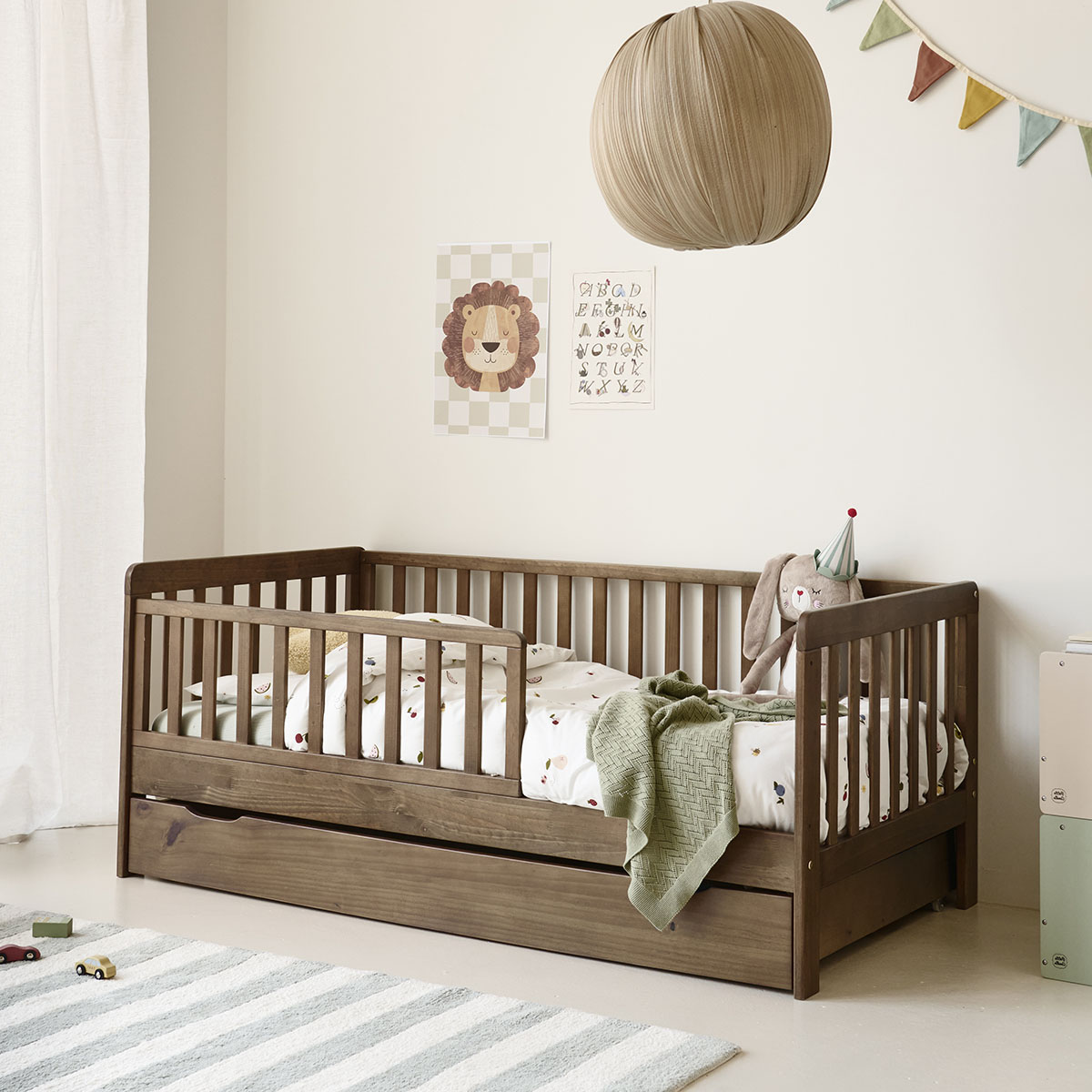Lit enfant Plume en bois foncé avec barrière de sécurité et tiroir de rangement, décoré avec du linge de lit imprimé, un doudou dans une chambre au style doux et naturel